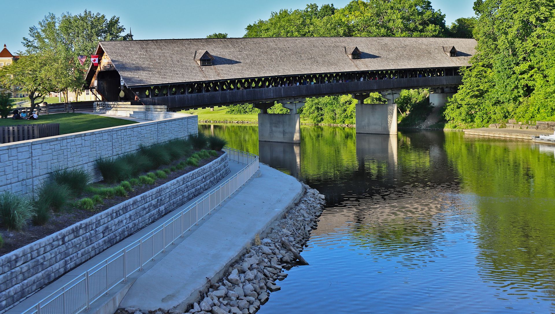 Thatched-roof bridge over a calm canal, with stone embankments and green trees in sunlight