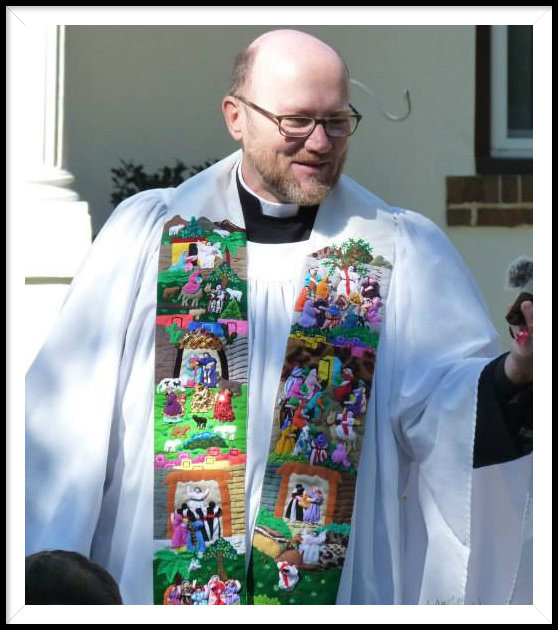 A smiling clergyman wears a white alb and a colorful stole embroidered with biblical scenes, standing outdoors.