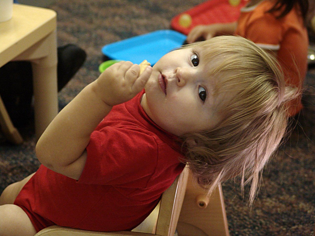 A child in a red shirt sits in a chair, looking back over their shoulder while holding a small piece of food.