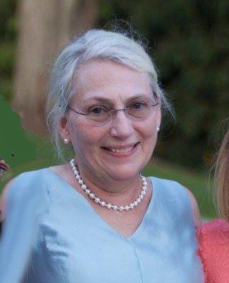 A smiling person with gray hair, wearing glasses, a pearl necklace, and a light blue dress, outdoors against greenery.