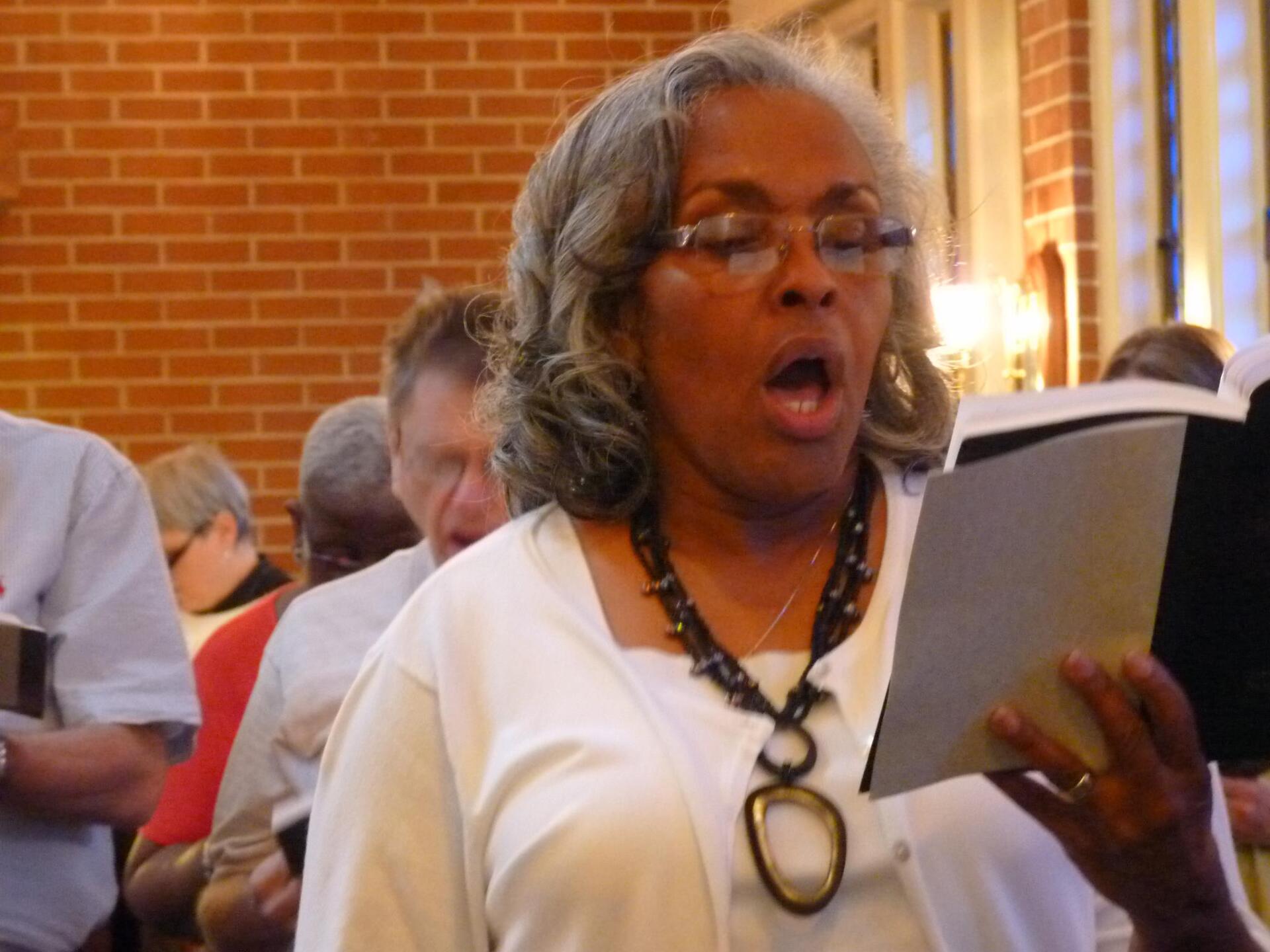 A person singing while holding an open hymnal in a choir, with a brick wall in the background.