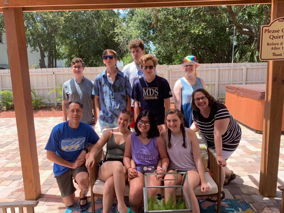 A group of eleven people gather for a photo on a patio under a wooden trellis, with a white fence in the background.