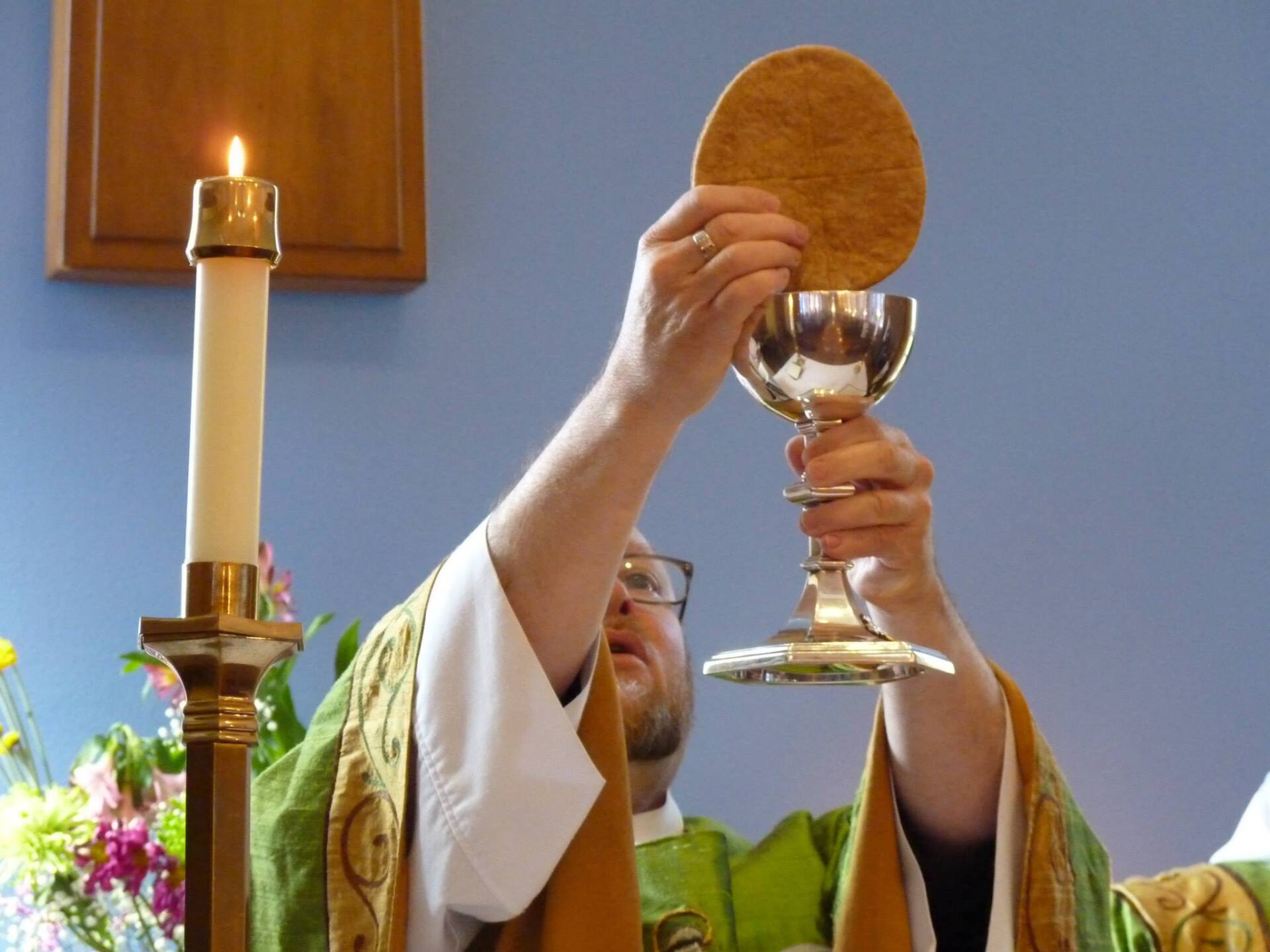 A priest in green liturgical vestments holds up a communion host above a chalice during a church service.