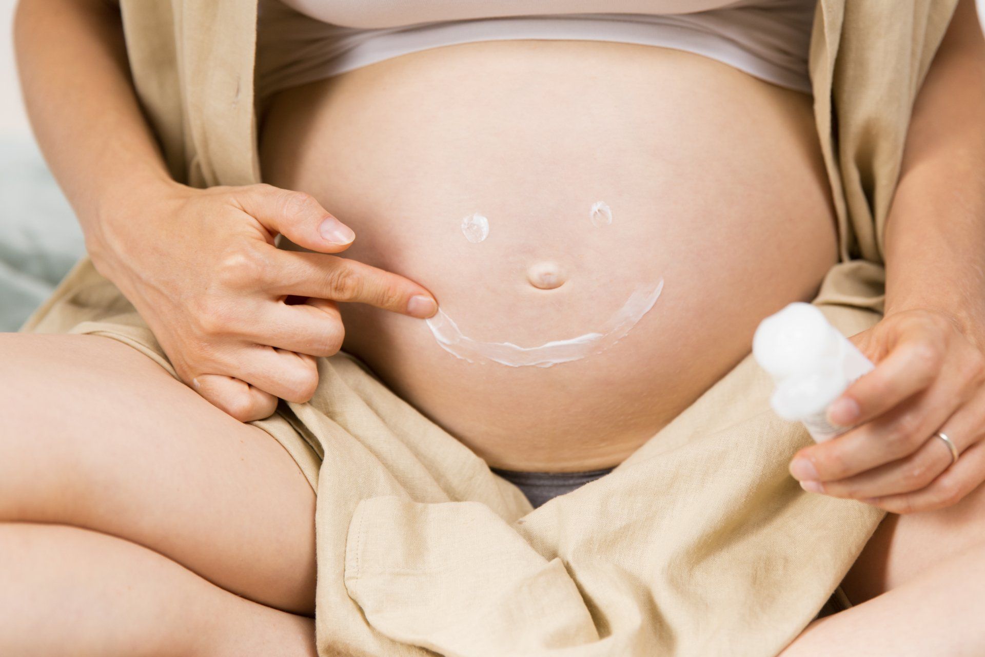 Mulher grávida aplicando loção na barriga, formando um sorriso
