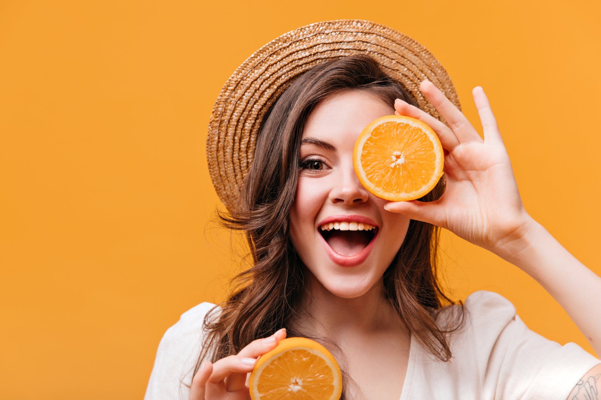 Mulher jovem usando chapéu, segurando uma fatia de laranja sobre o rosto