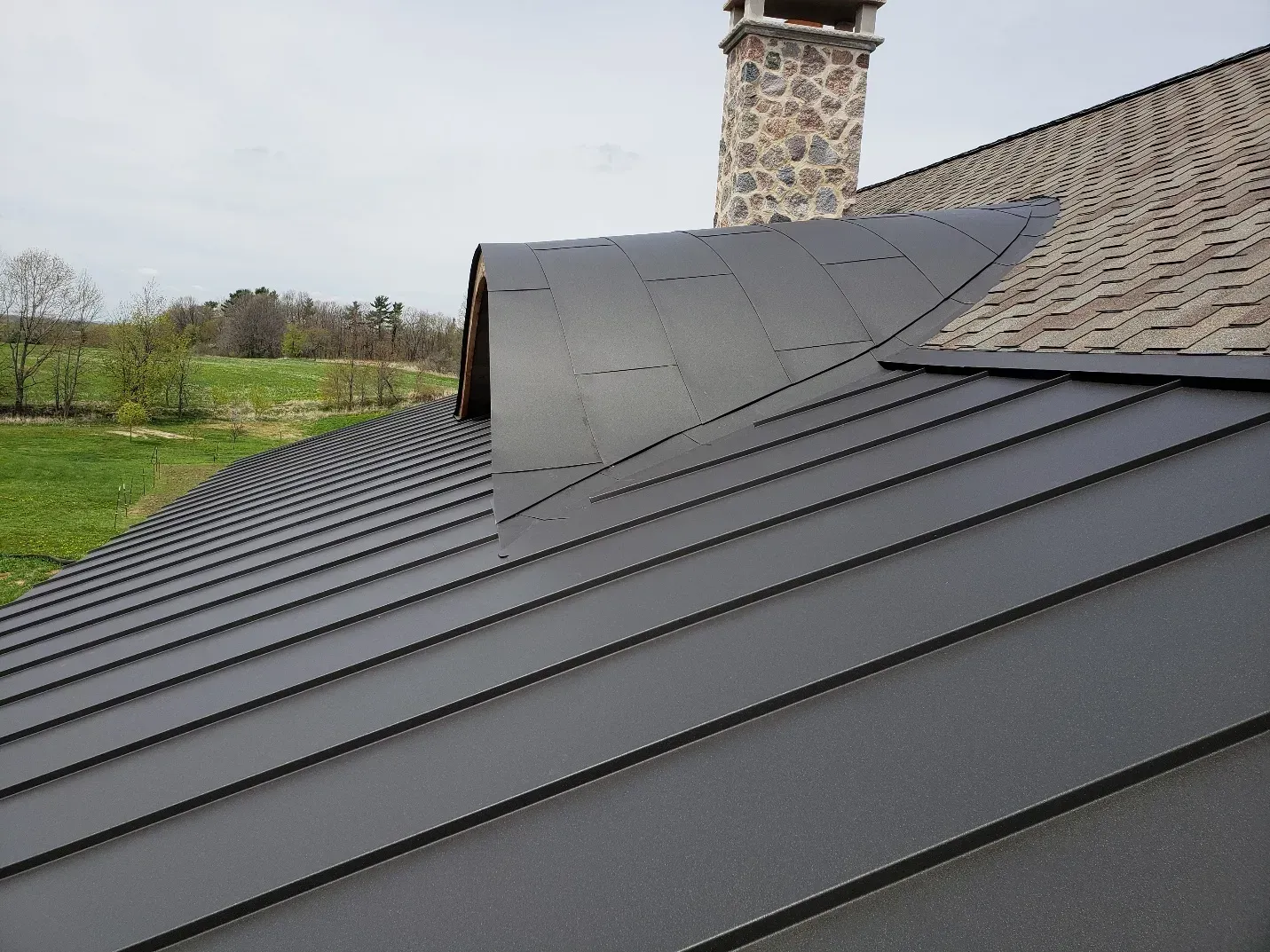 Close-up detail of curved standing seam metal roof panels near a stone chimney in Sheboygan, WI