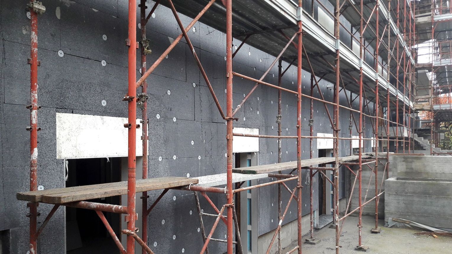 Facciata di un edificio con impalcature, pareti grigie e supporti metallici rossi, in un cantiere edile.