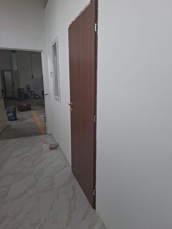 Corridoio interno con pareti bianche, porta marrone e pavimento piastrellato che conduce a una stanza in penombra.