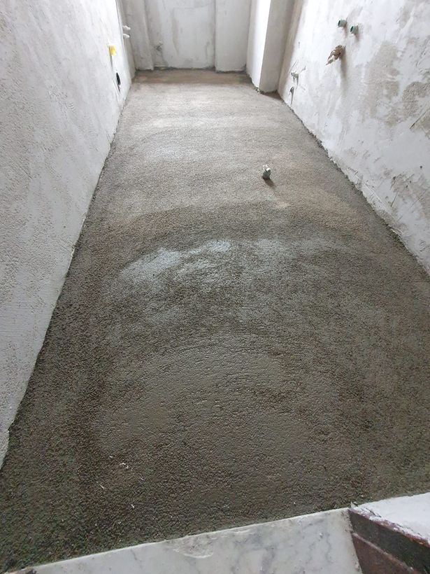 Stretto corridoio in cemento grezzo con pareti bianche e pavimento grigio ruvido.
