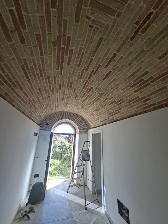 Stretto corridoio con soffitto a volta in mattoni, pareti bianche e una scala vicino a una porta illuminata dal sole.
