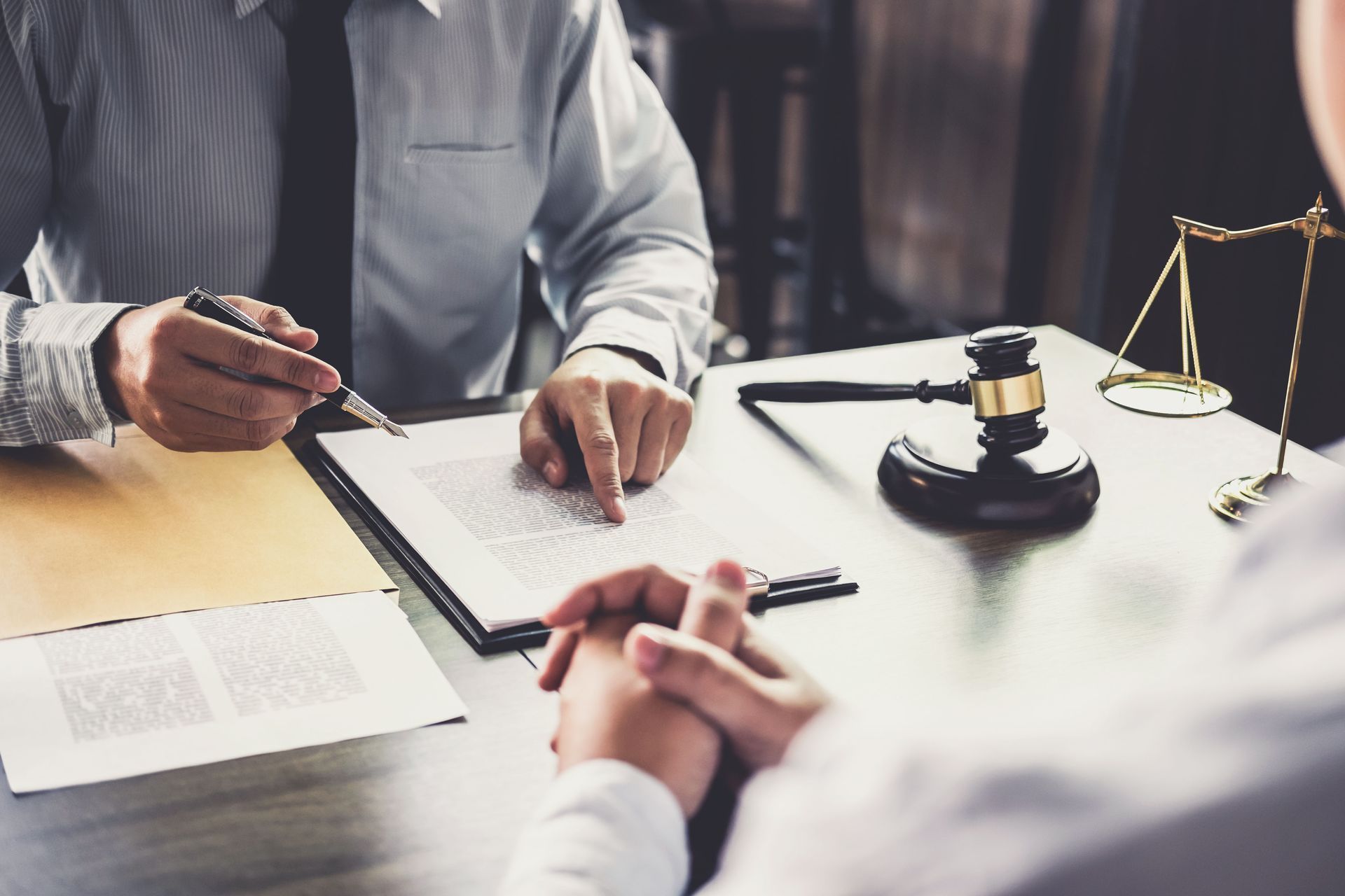 A lawyer in a dress shirt points to a document across a desk from a client, with a judge’s gavel and scales of justice.