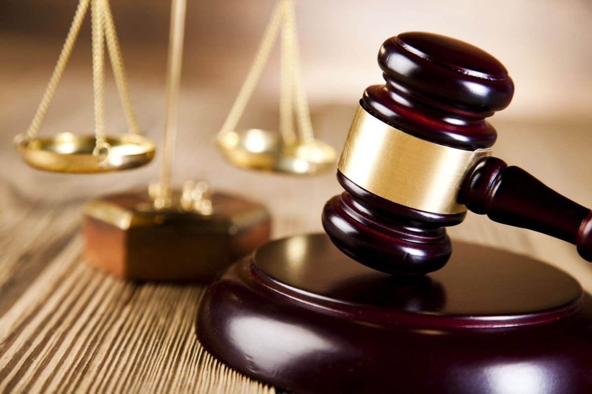 A polished wooden gavel rests on its block in the foreground with out-of-focus golden scales of justice in the back.