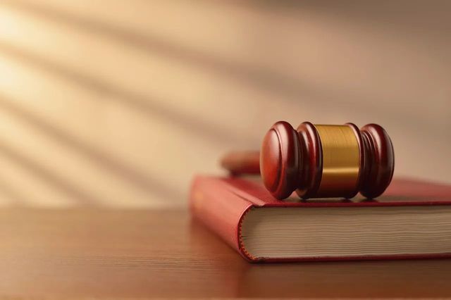 A polished wooden gavel with a gold band rests on top of a closed red book against a soft-lit, neutral background.