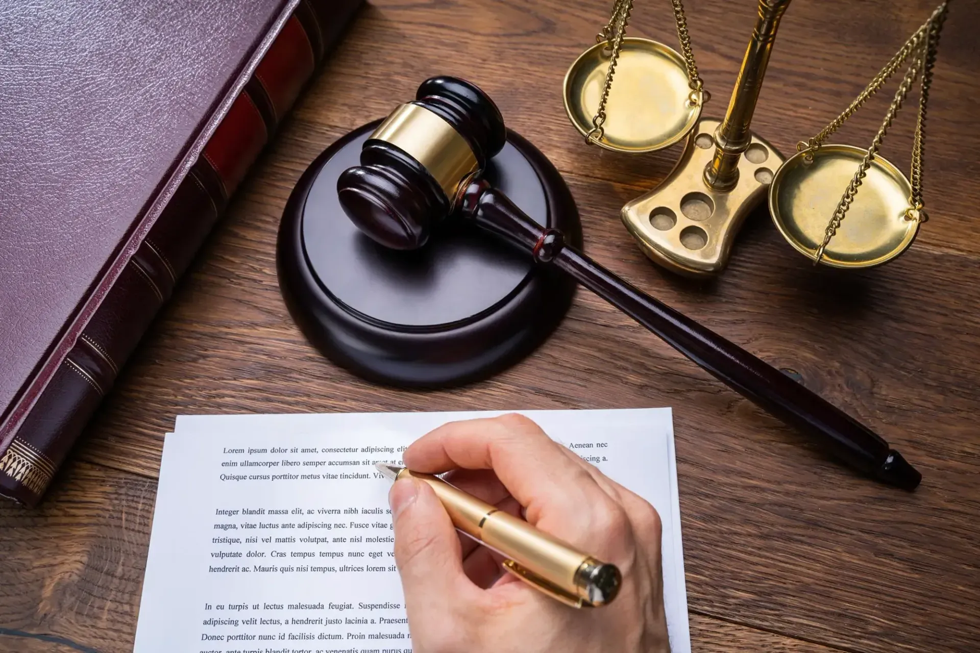 A hand signs a document on a desk next to a gavel, a legal book, and a gold scale of justice.