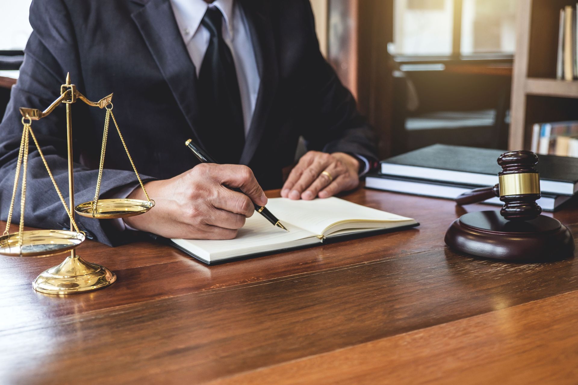 A person in a suit writes in a notebook at a wooden desk with a gold scale of justice and a wooden gavel.