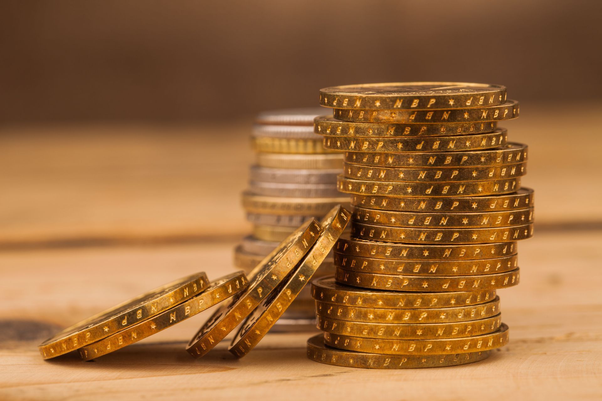 A tall stack of gold coins sits on a wooden surface next to a smaller, slanted pile of gold coins.
