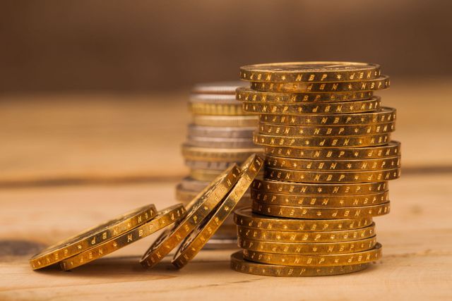 A tall stack of gold coins sits on a wooden surface next to a smaller, slanted pile of gold coins.