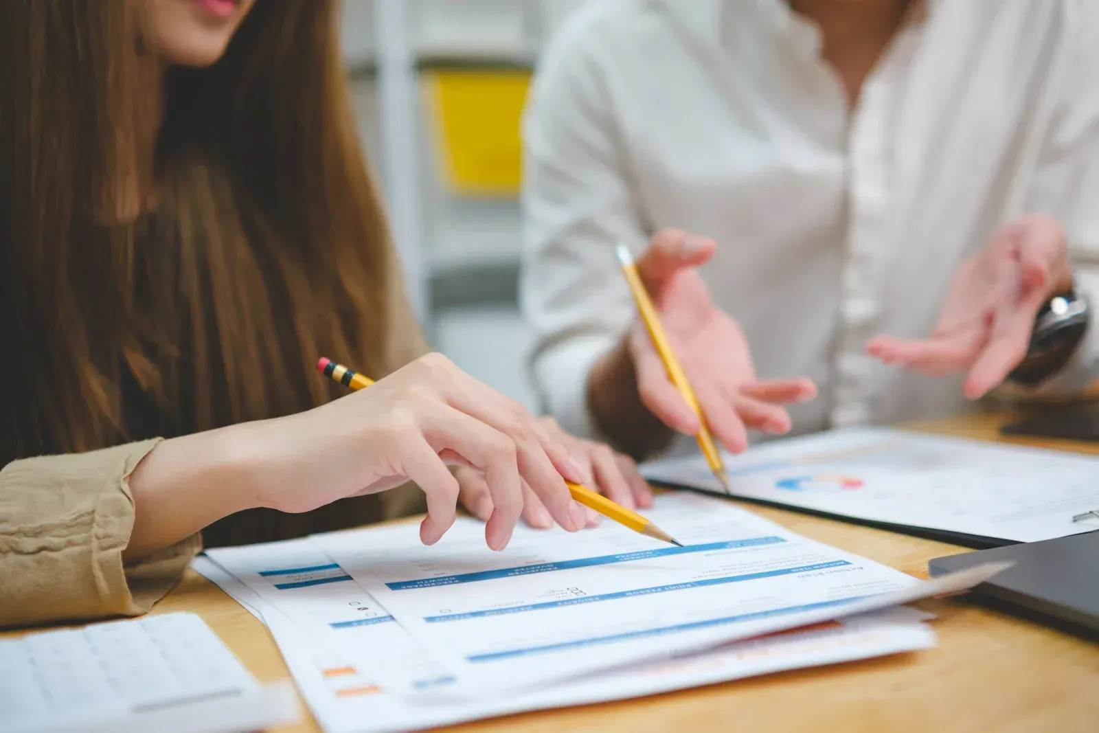 Two people collaborate at a wooden desk, analyzing business charts and spreadsheets while holding pencils.