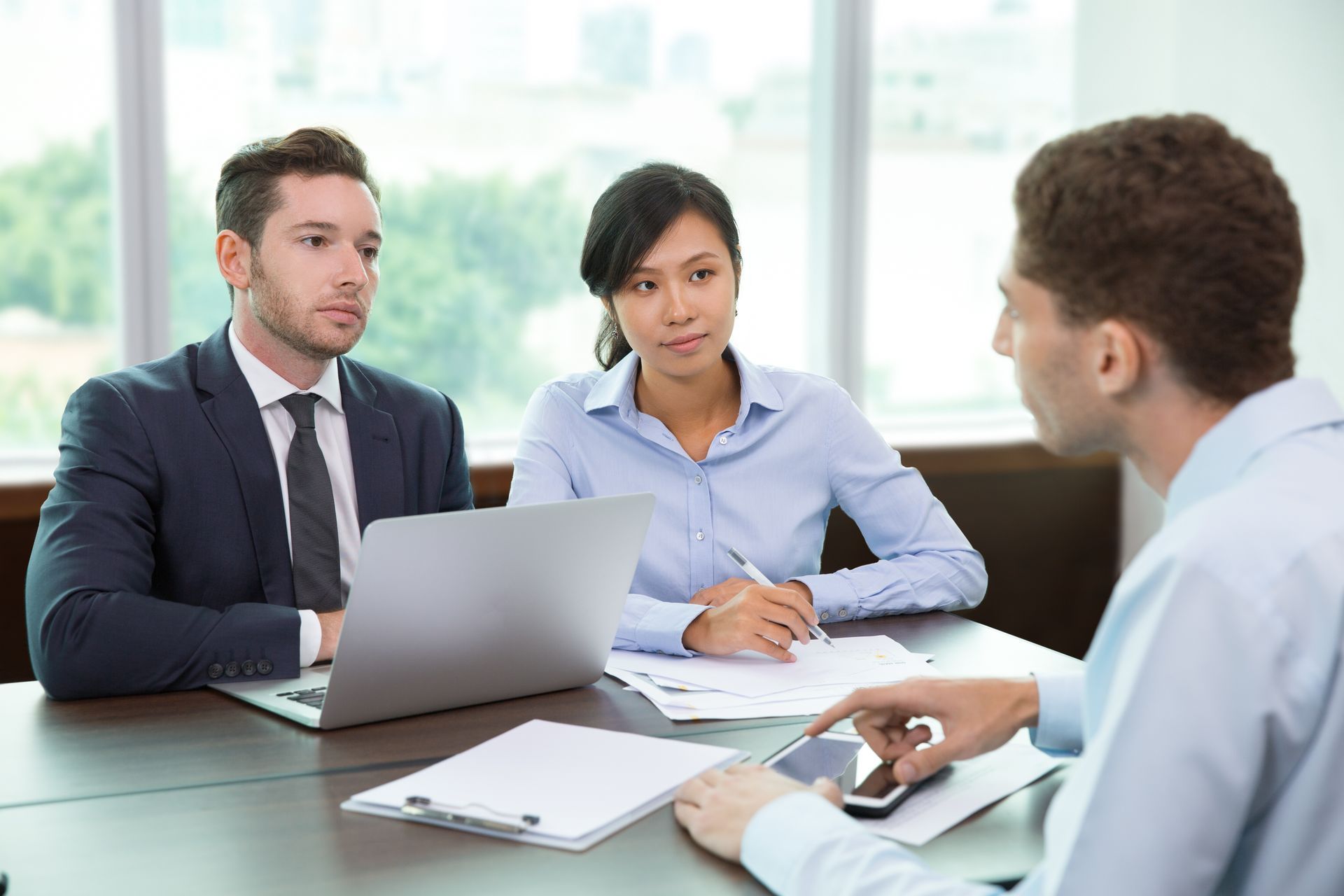 Three people sit around a desk in an office, engaged in a professional meeting or job interview.