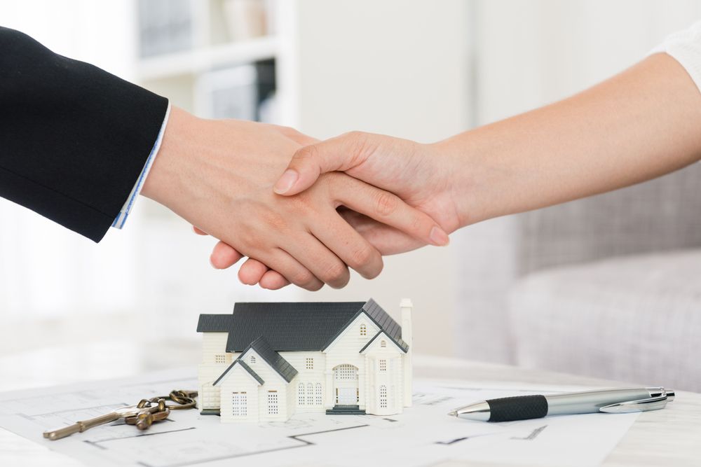 Two People Shaking Hands Over a Miniature House — Kellie O'Malley Conveyancing In Lake Macquarie, NSW