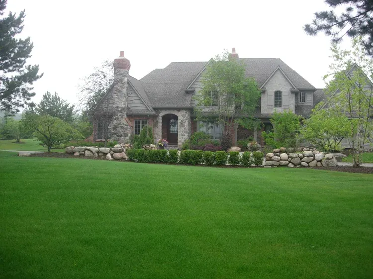Stone house with arched entry and large green lawn, surrounded by shrubs and trees