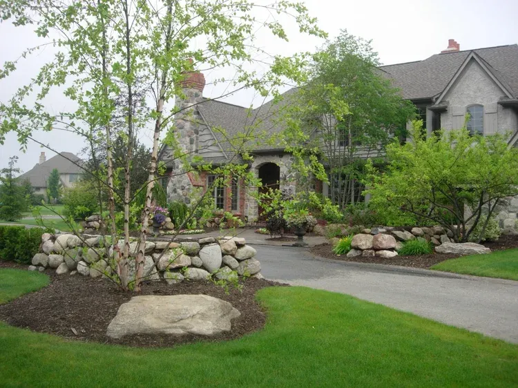 Stone-lined front garden with curved walkway leading to a gray house and trees