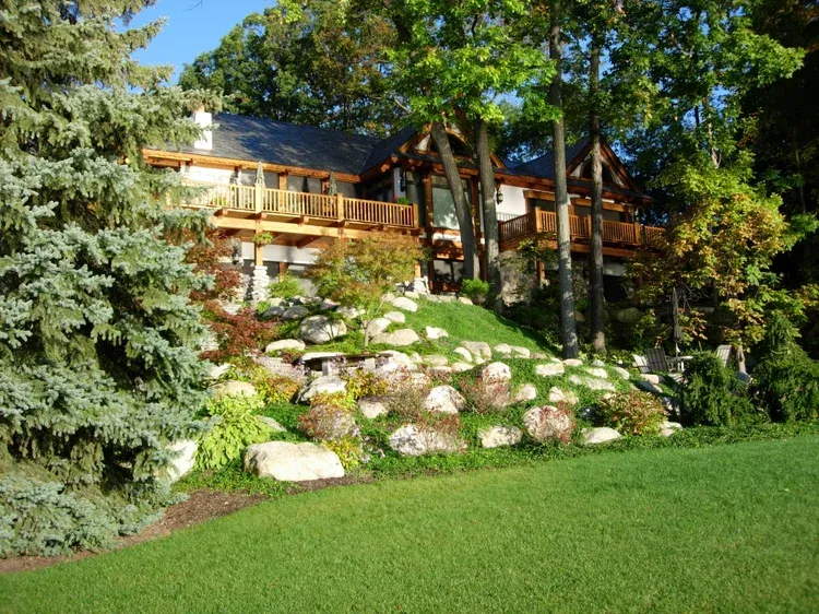 Wooden house on a sunny hillside with trees, rocks, and a green lawn