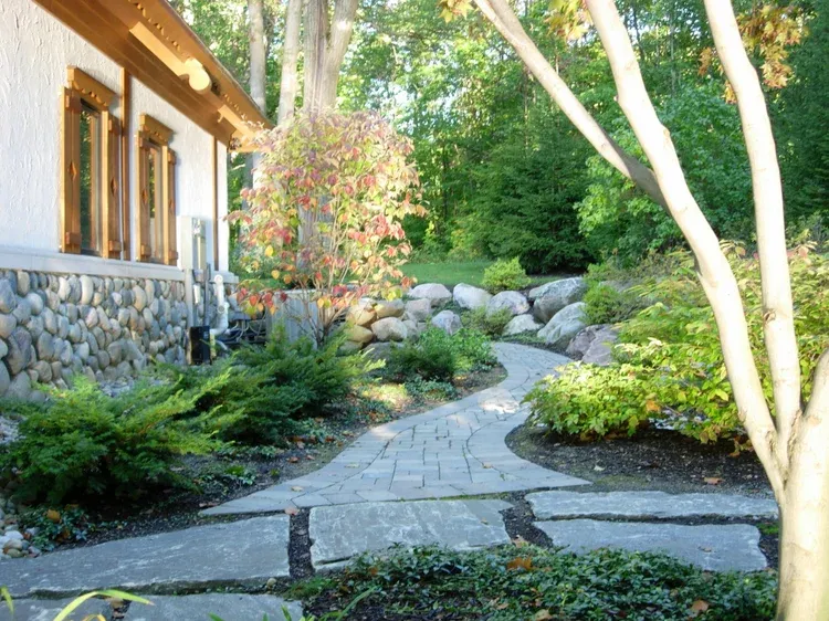 Stone garden path beside a house, lined with shrubs, rocks, and sunlit trees