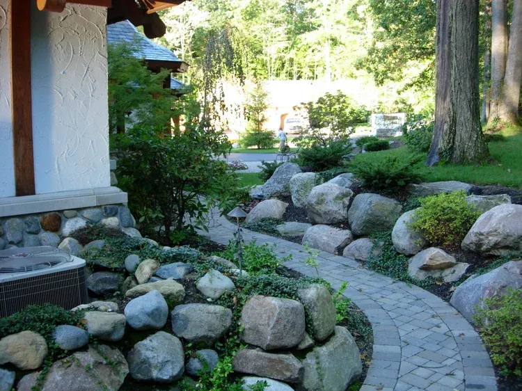 Stone garden path beside a house, lined with rocks and greenery in a wooded yard