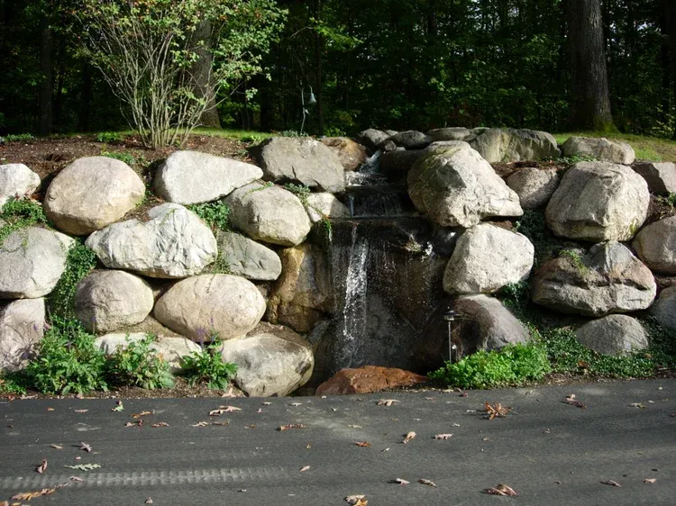 Stone retaining wall with a small waterfall spilling onto pavement in a wooded area