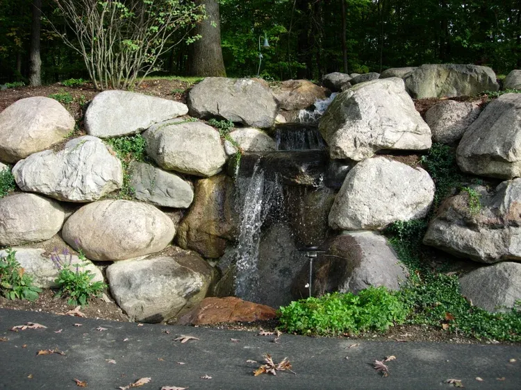 Stone wall fountain with water flowing into a small basin beside a paved path and greenery