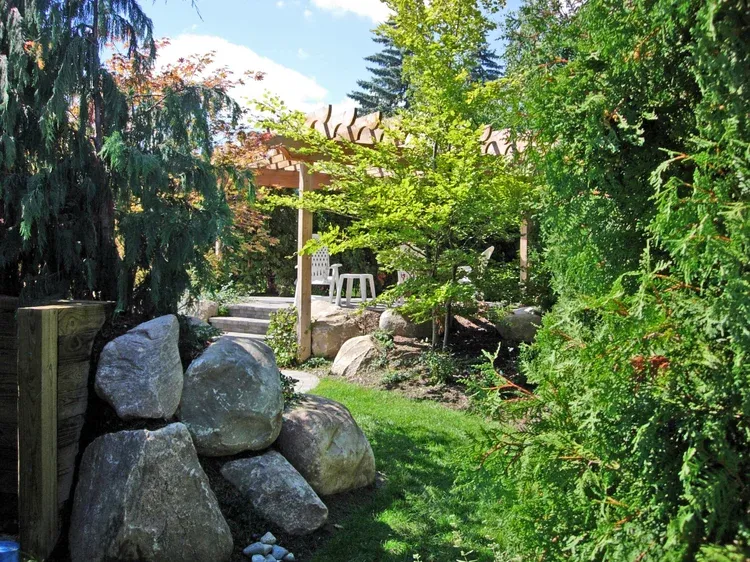 Garden path with rocks, lush green trees, and a shaded patio in the background