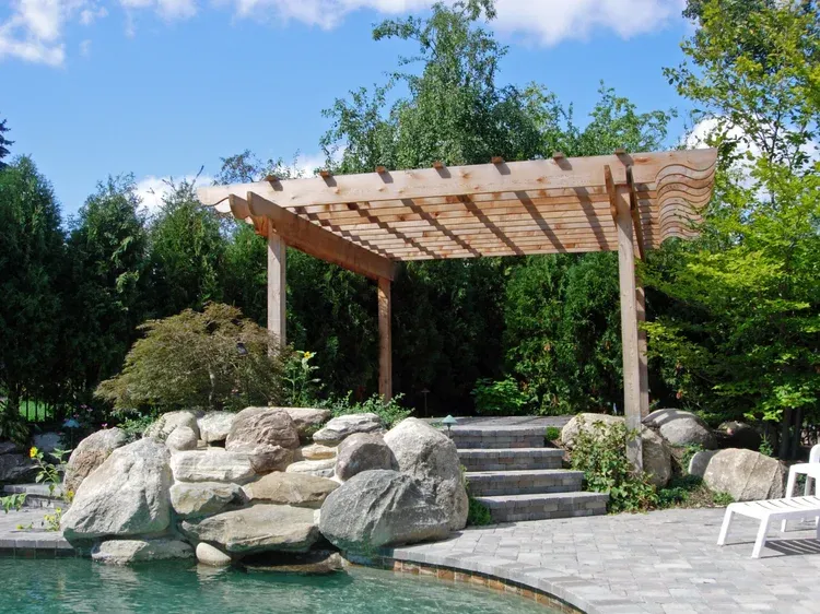 Backyard patio with wooden pergola, stone steps, rocks, and a pool under a blue sky