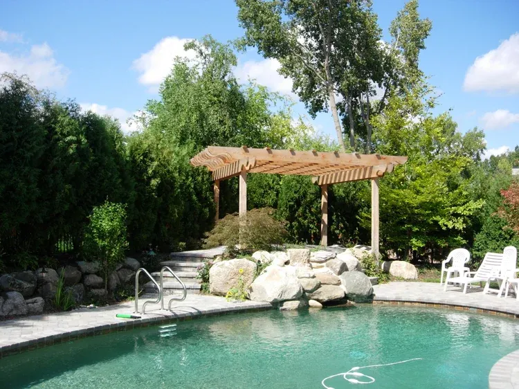 Backyard pool with stone landscaping, white lounge chairs, and a wooden pergola surrounded by trees