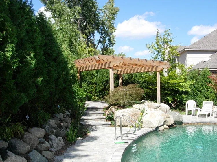 Backyard pool with stone path, wooden pergola, and white chairs beside lush greenery