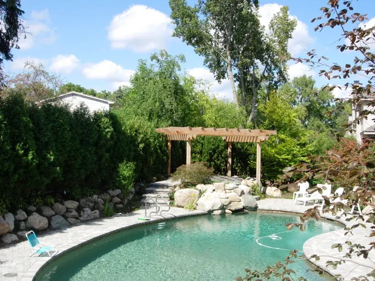 Backyard pool with stone patio, pergola, and lush green trees under a blue sky
