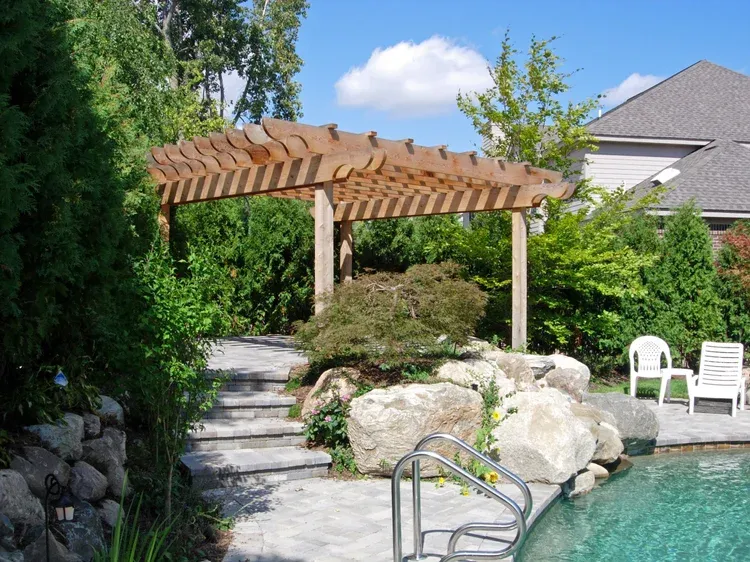 Backyard pool with stone path, pergola, white chairs, and lush greenery
