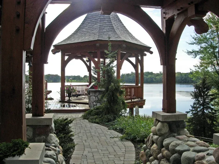 Gazebo and stone walkway overlooking a calm lake, framed by wooden arches and potted plants