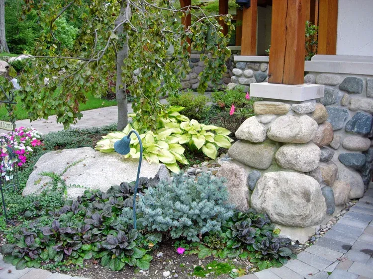 Landscaped garden bed with green and blue plants, stone edging, and a wooden porch pillar