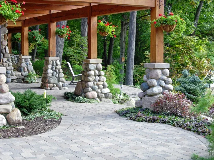 Stone-pillared wooden garden pergola with hanging flower baskets and a curved brick path amid lush greenery