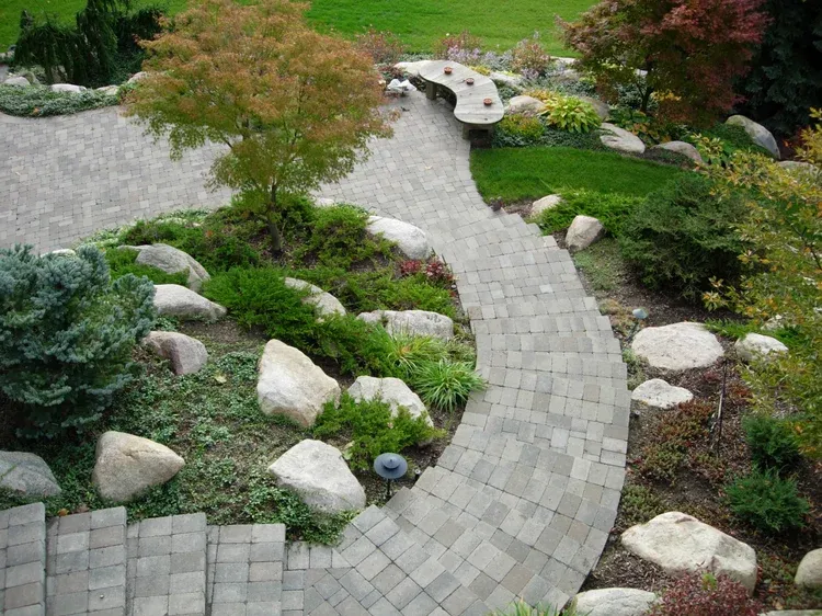 Curved stone path through a landscaped garden with rocks, shrubs, and a small wooden bridge