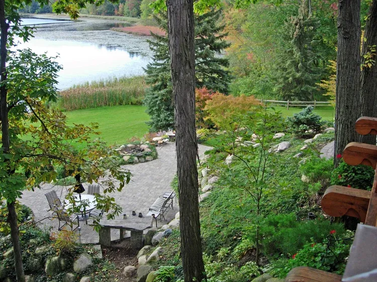 Lakeside garden with stone patio, lawn, trees, and a winding path beside calm water