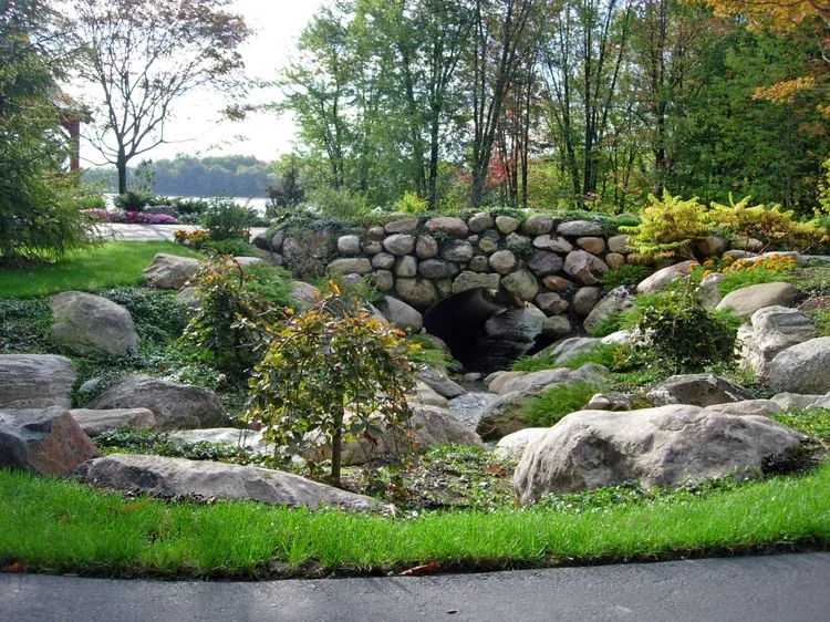 Landscaped garden with a stone arch, lush greenery, rocks, and a curved paved path.