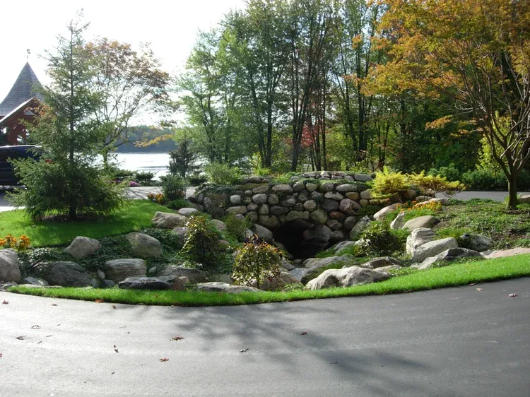Stone garden bridge over a landscaped stream, with green trees and a road in the foreground