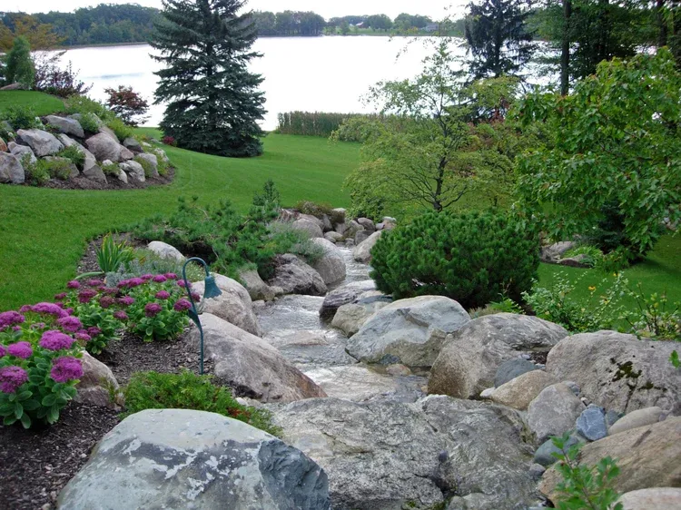 Rock garden stream with large stones, purple flowers, and a lake in the background