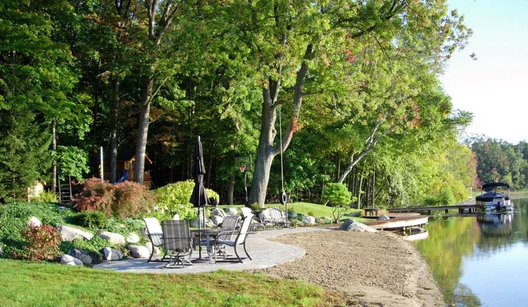 Lakeside patio with white chairs, green trees, and a calm shoreline beside a dock