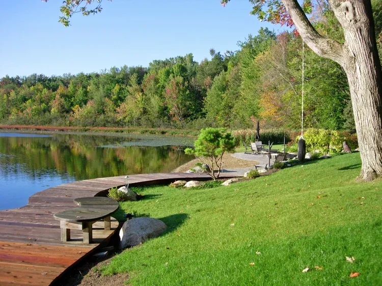 Lakeside dock with green lawn, round tables, and autumn trees reflected in calm water