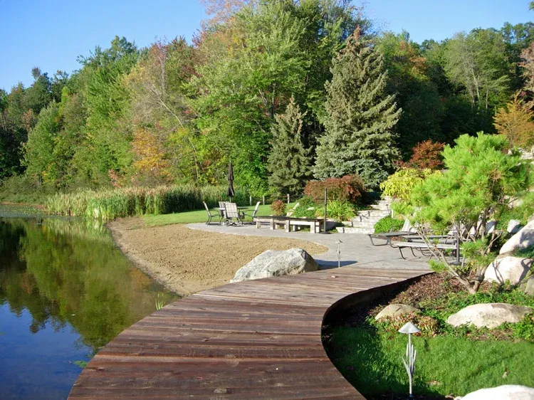 Curved wooden boardwalk by a calm lake with lounge chairs, rocks, and green trees in autumn light