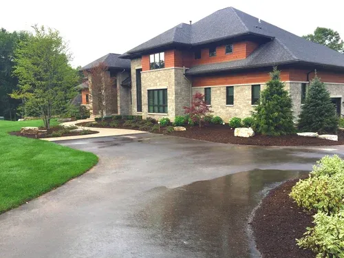 Large stone-and-wood house with a wet driveway and landscaped front yard after rain