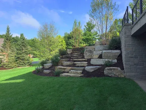 Stone steps in a landscaped backyard beside a green lawn and trees under a blue sky.